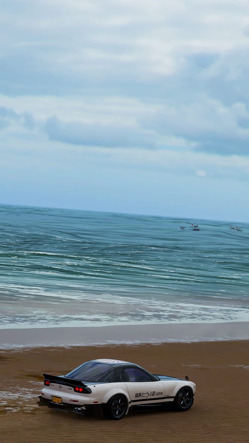 White Mazda RX-7 sports car parked on a sandy beach with rolling ocean waves and a cloudy sky in the background.