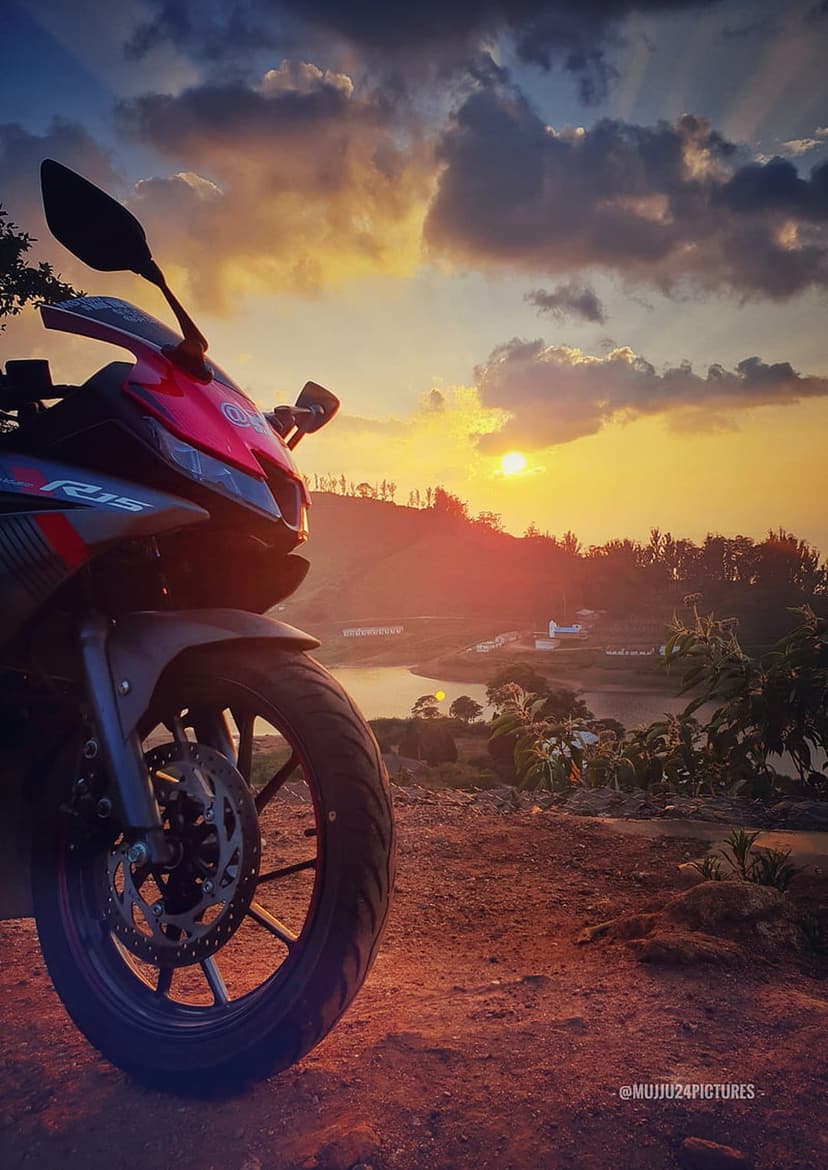 Red and black sport motorcycle parked on a dirt path facing a calm lake under a dramatic orange sunset sky.