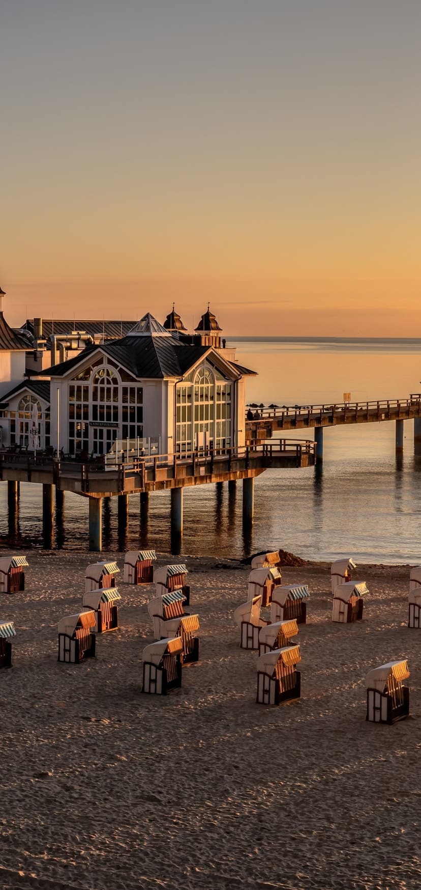 Sellin Pier's white building stands over the ocean during sunrise with beach chairs on the sand below.