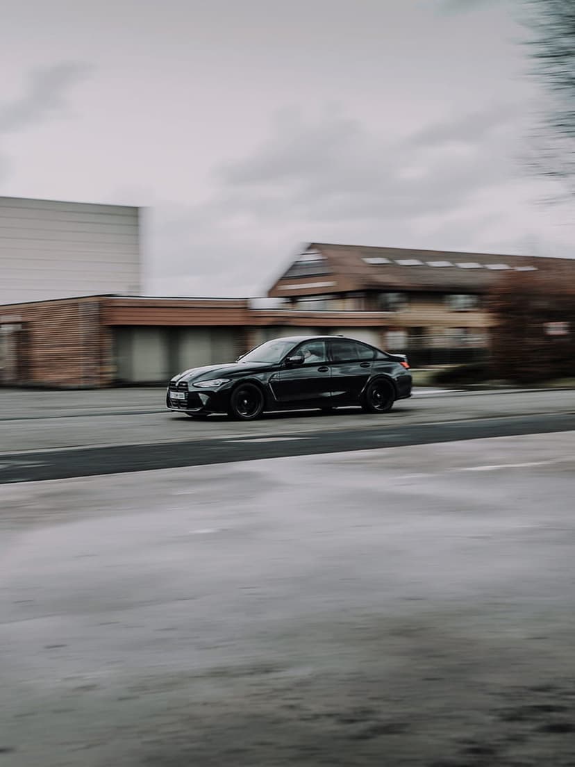 Black BMW M3 sports car speeds along a rain-slicked road featuring heavy motion blur and moody industrial scenery.