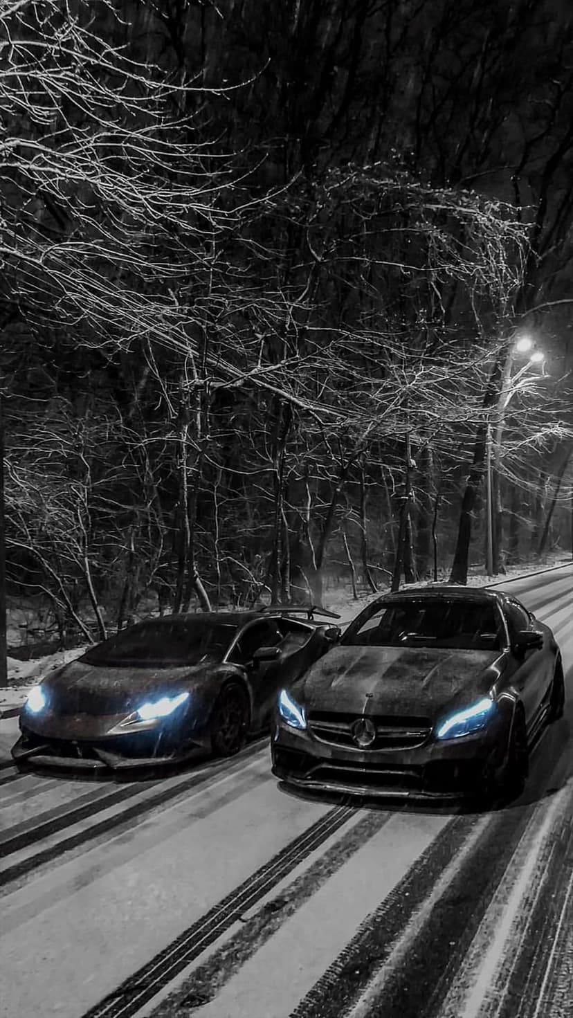 Lamborghini and Mercedes-Benz sports cars parked on a snowy forest road at night with glowing headlights.