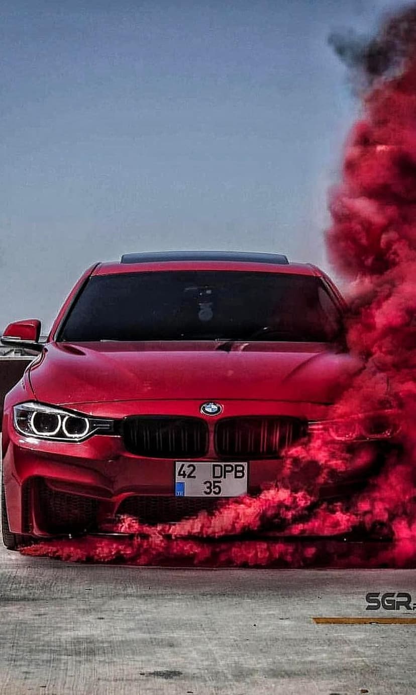 Red BMW with glowing headlights stands before a clear blue sky as thick clouds of red smoke surround the chassis.