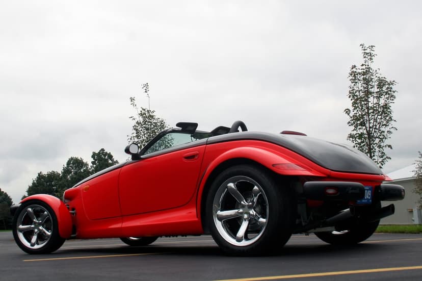 Red and black Plymouth Prowler with chrome wheels from a low rear angle on an asphalt lot under a cloudy sky.