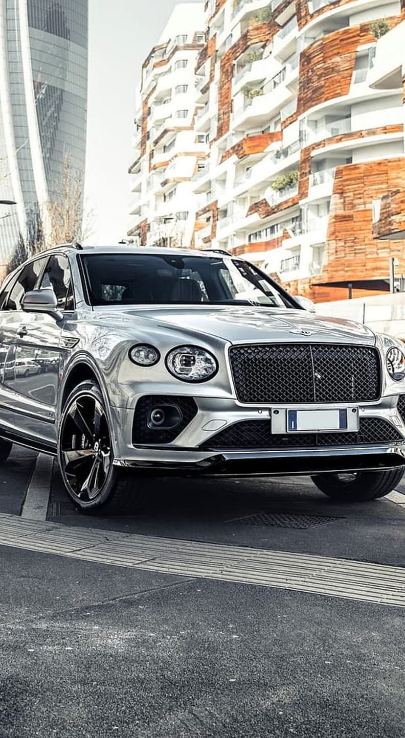 Silver Bentley Bentayga SUV parked on an urban street against a backdrop of modern geometric building facades.