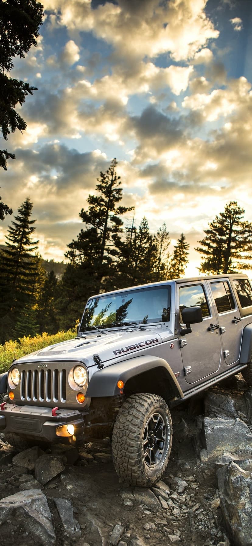 Silver Jeep Wrangler Rubicon parked on rugged rocks under a golden sunset sky with pine trees in the background.