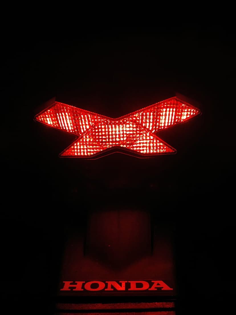 Red X-shaped motorcycle tail light with a glowing Honda logo beneath it against a dark, low-light background.