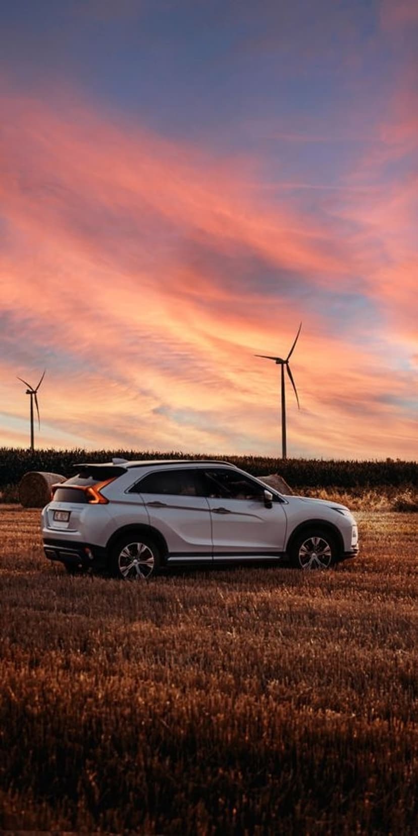 White SUV parked in a wheat field at sunset with wind turbines and hay bales under a vibrant orange and pink sky.