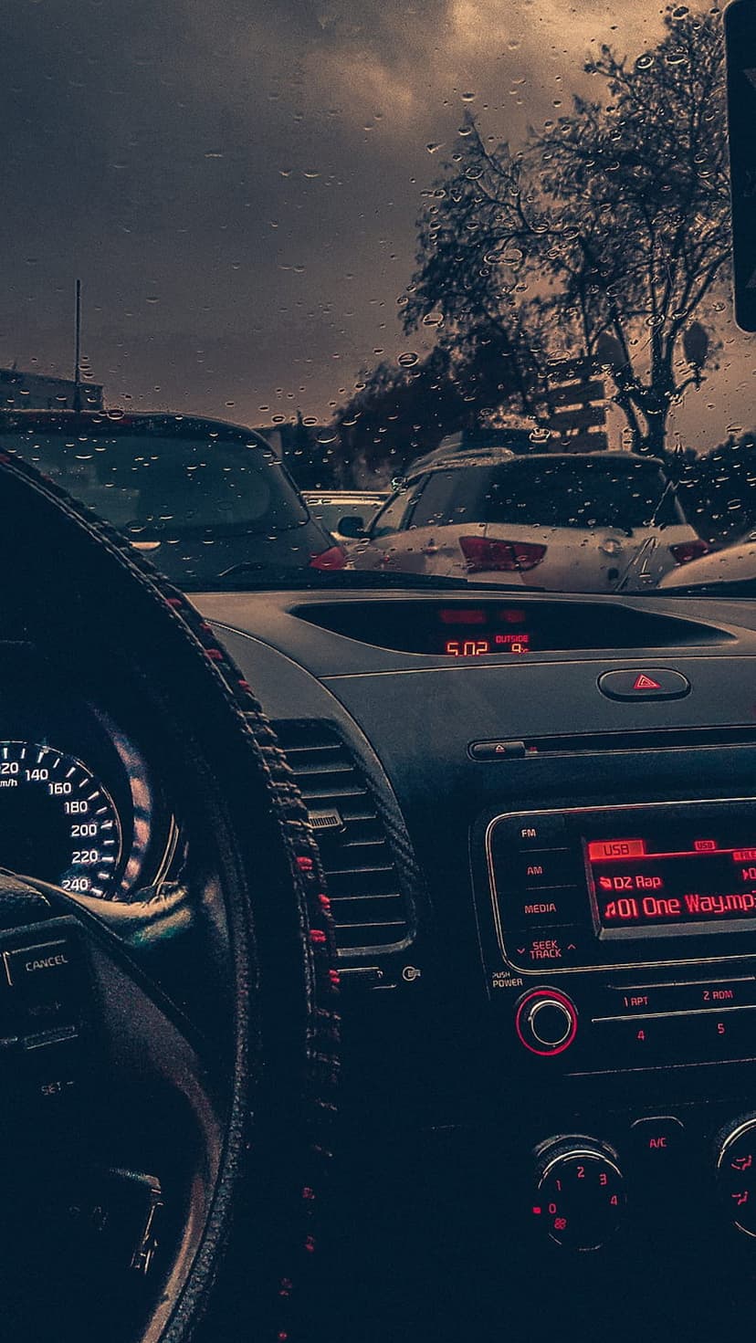 Car dashboard with red glowing instruments faces a rainy windshield blurred with city lights and distant trees.