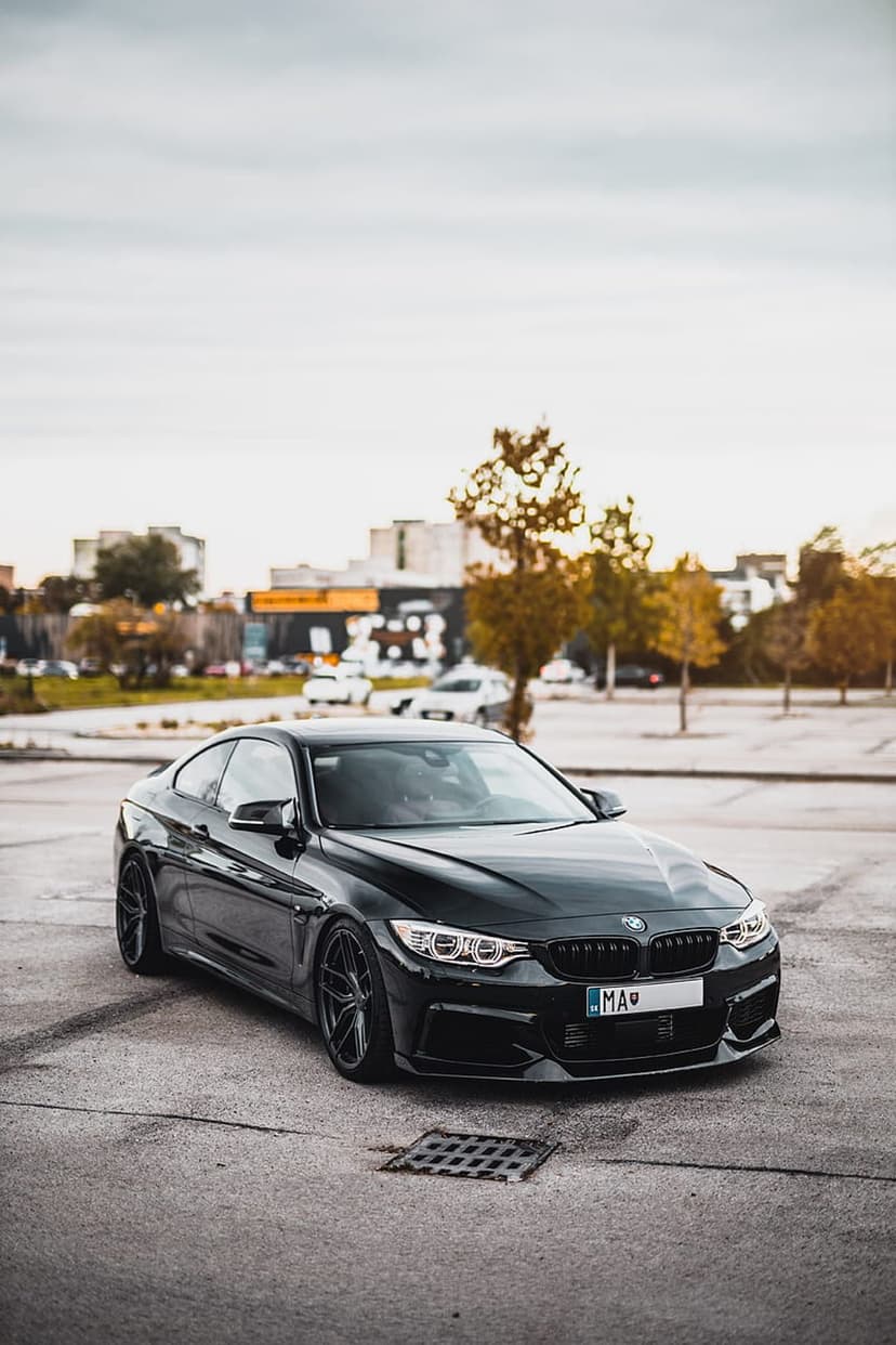 Black BMW coupe with glowing headlights parked on asphalt against a cloudy city skyline background.