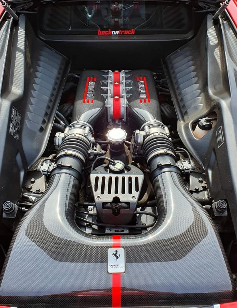 Ferrari engine bay with carbon fiber texture, red valve covers, and silver prancing horse logo on the manifold.