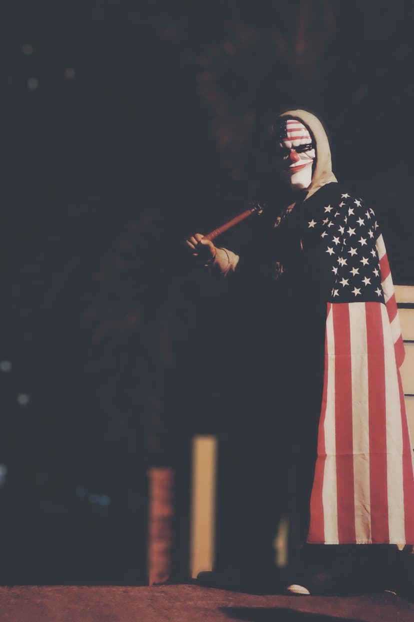 Masked person wearing an American flag cape and hoodie holds a reddish rod against a dark, grainy background.