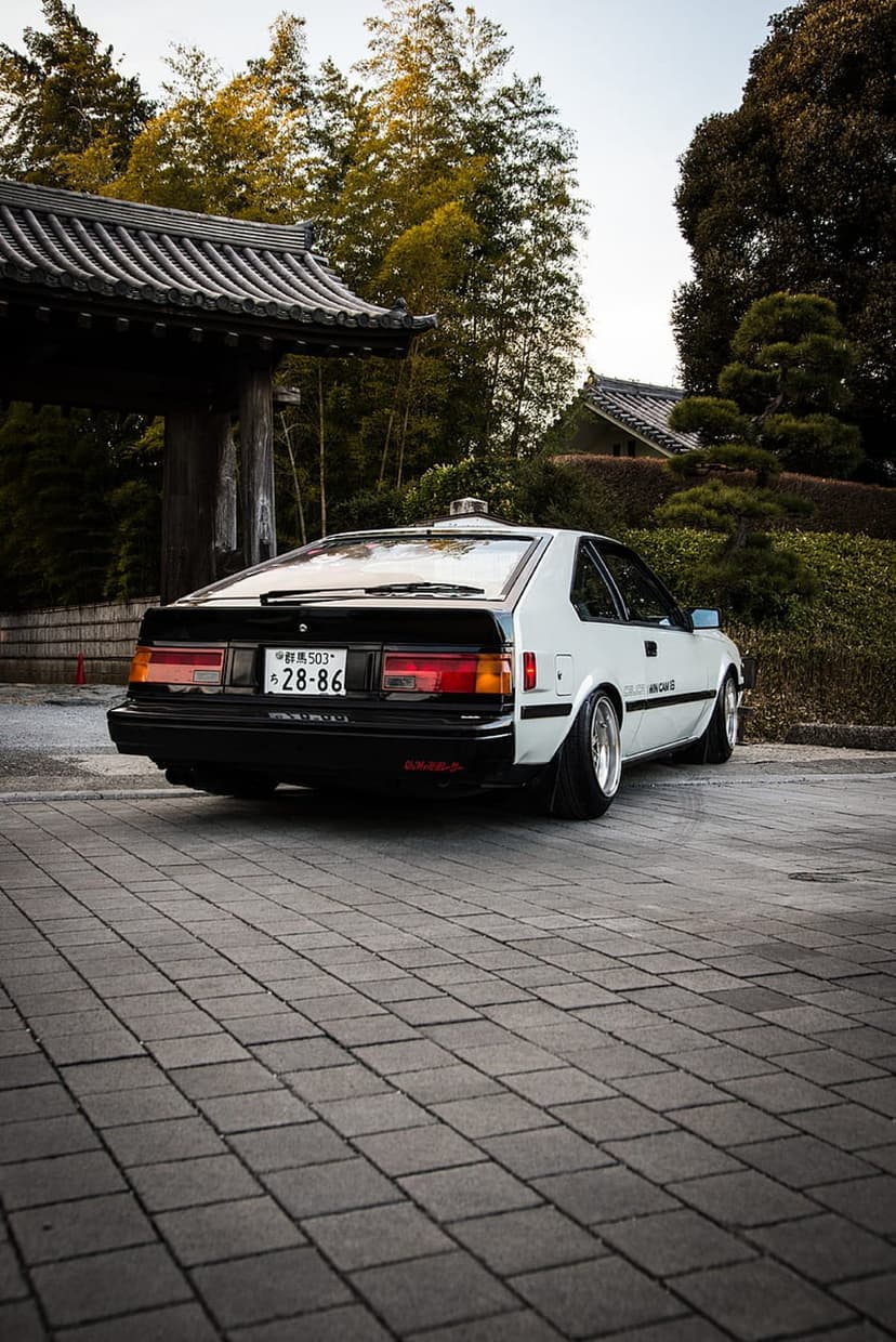 White Toyota Celica GT rear view parked on cobblestones with a traditional Japanese temple gate in the background.