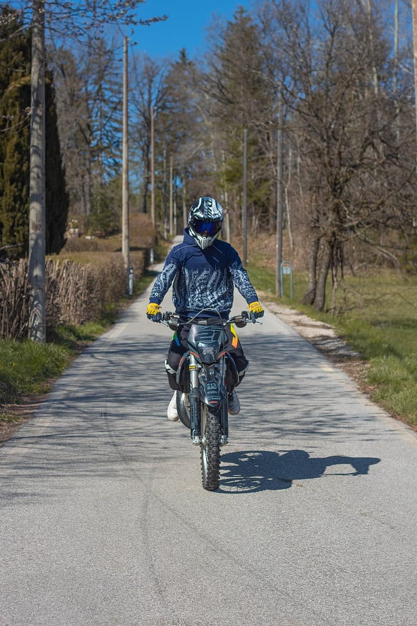 Motorcyclist in a blue hoodie and yellow gloves riding a dirt bike along a sunlit road lined with lush green trees.