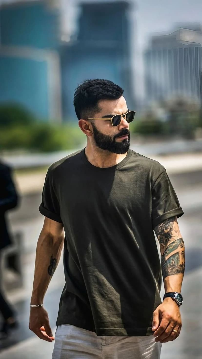 Virat Kohli wears dark sunglasses and an olive t-shirt while showcasing detailed arm tattoos in a city setting.