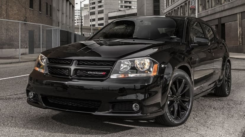 Front view of a glossy black Dodge Avenger sedan with black wheels on a city street with brick buildings.
