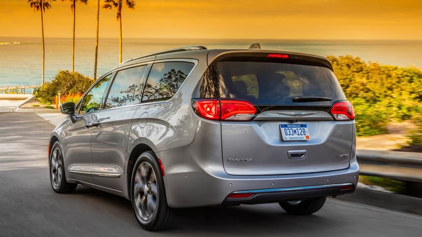 Silver Chrysler Pacifica minivan driving on a coastal road past palm trees under a golden sunset sky.