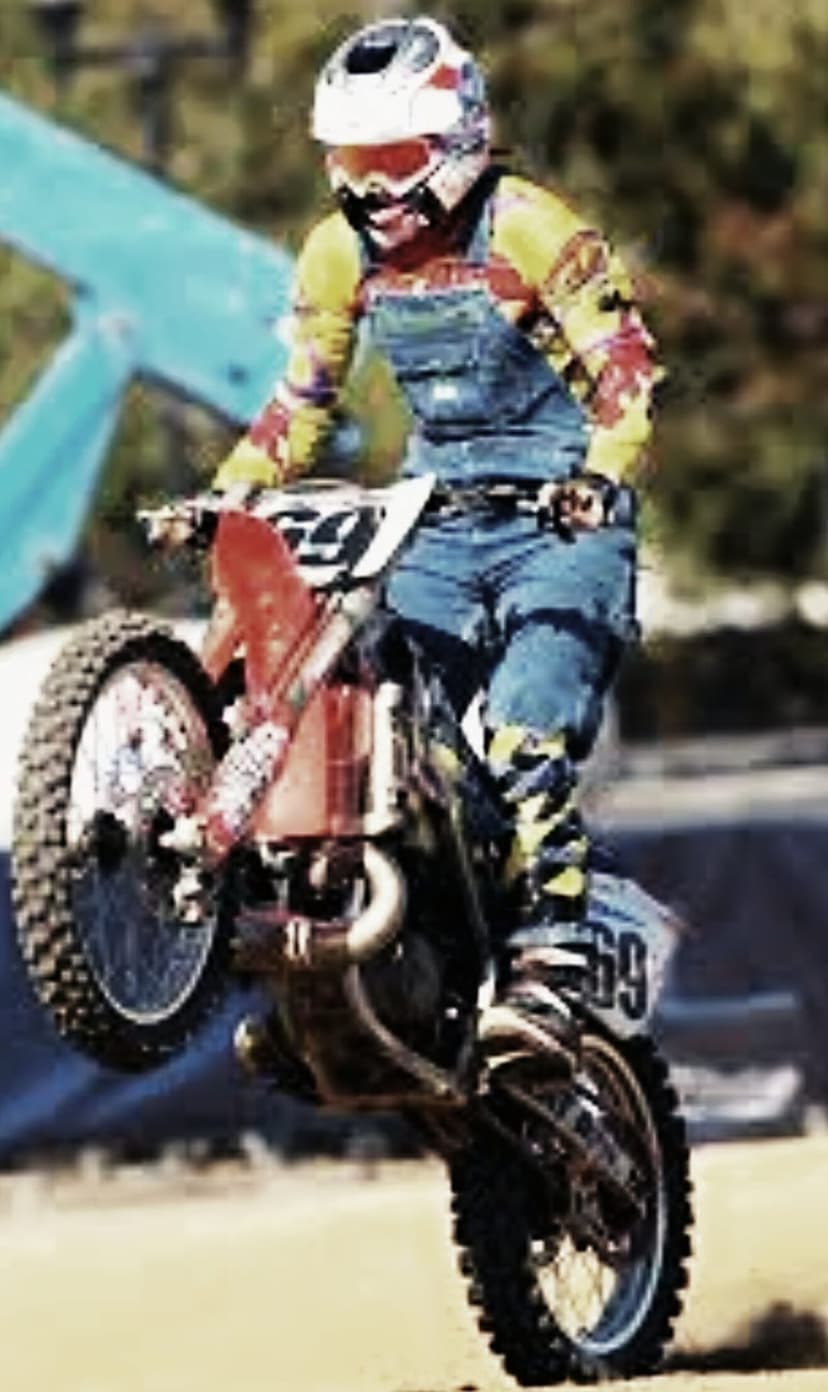 Motocross rider in denim overalls and white helmet performs a wheelie on a dirt bike with a red fender.
