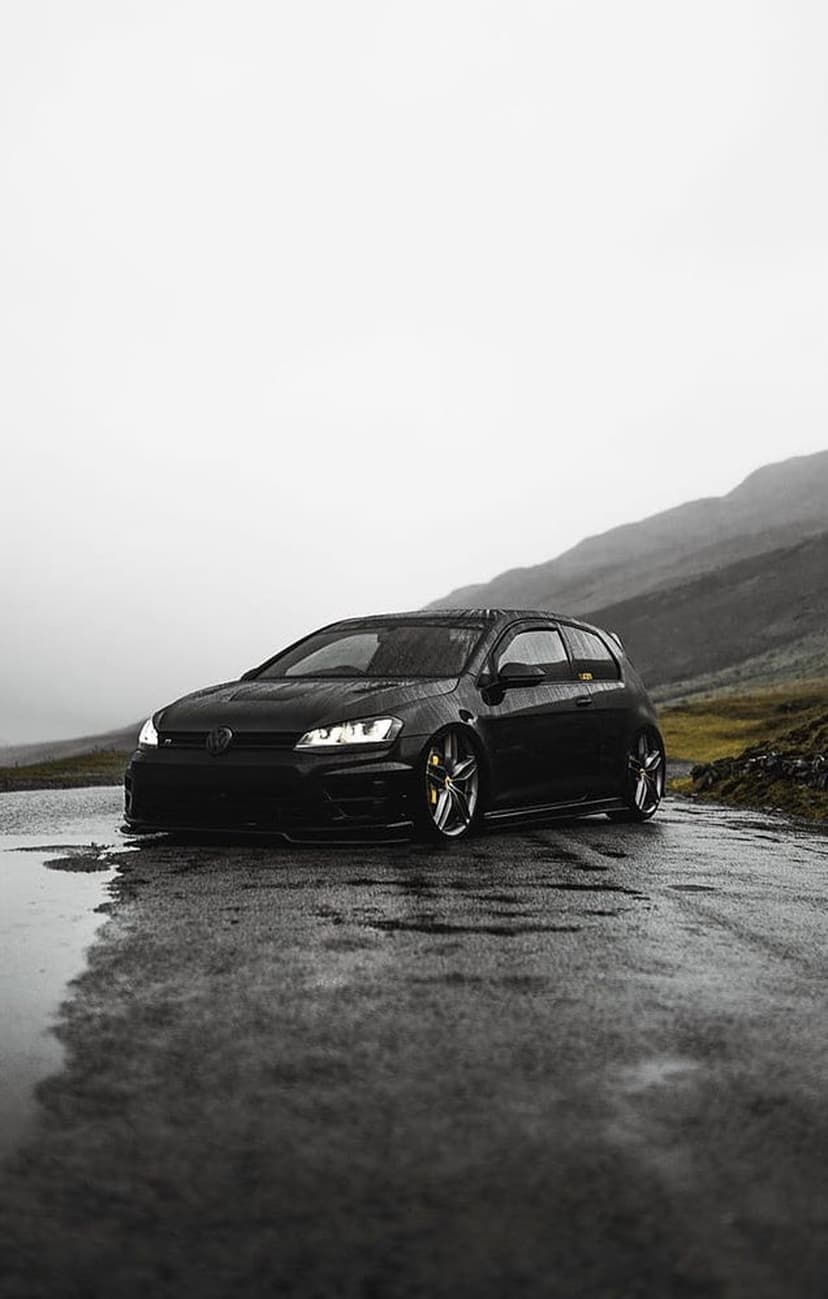 Black Volkswagen Golf R parked on a wet asphalt road with misty mountains and foggy skies in the background.