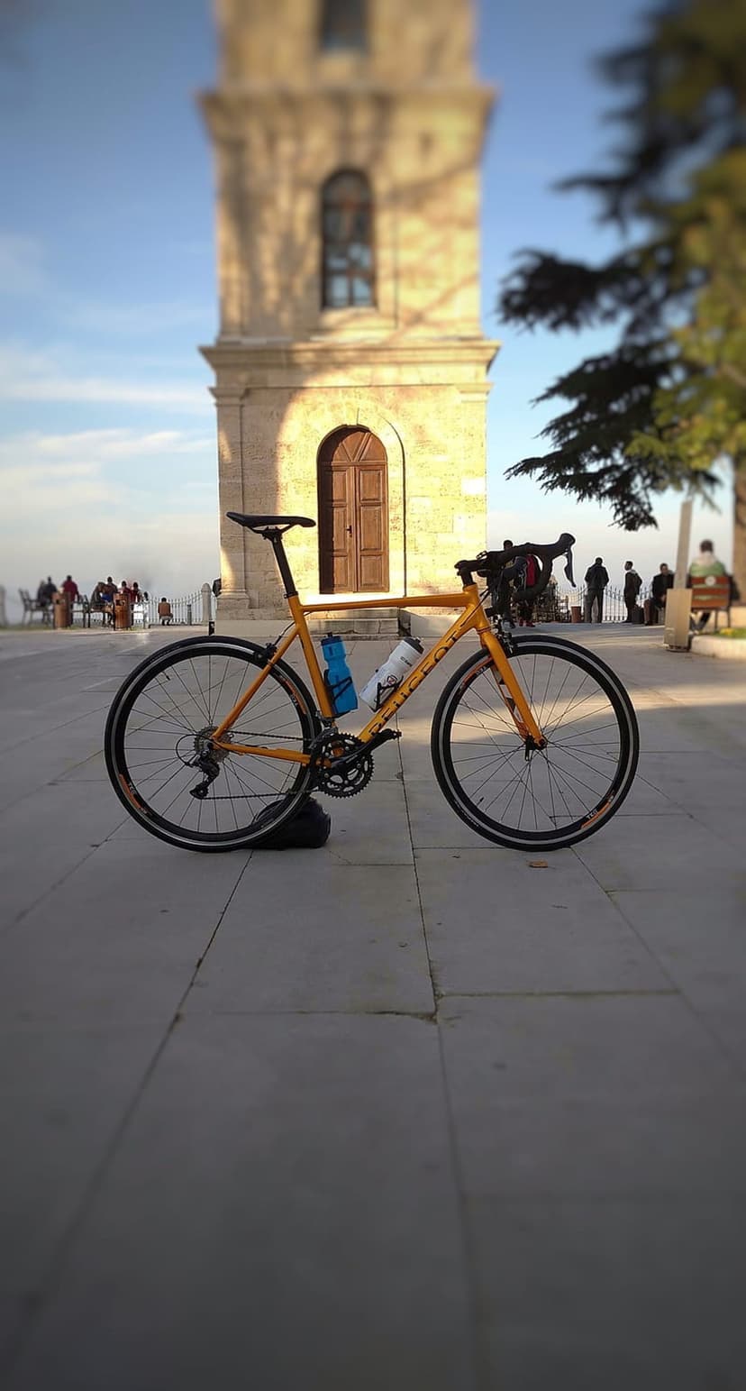 Orange bicycle parked on a stone plaza with a tall historic clock tower and green trees in the background.