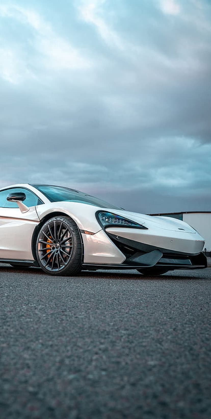 White McLaren supercar parked on dark asphalt with orange brake calipers under a dramatic cloudy sky.