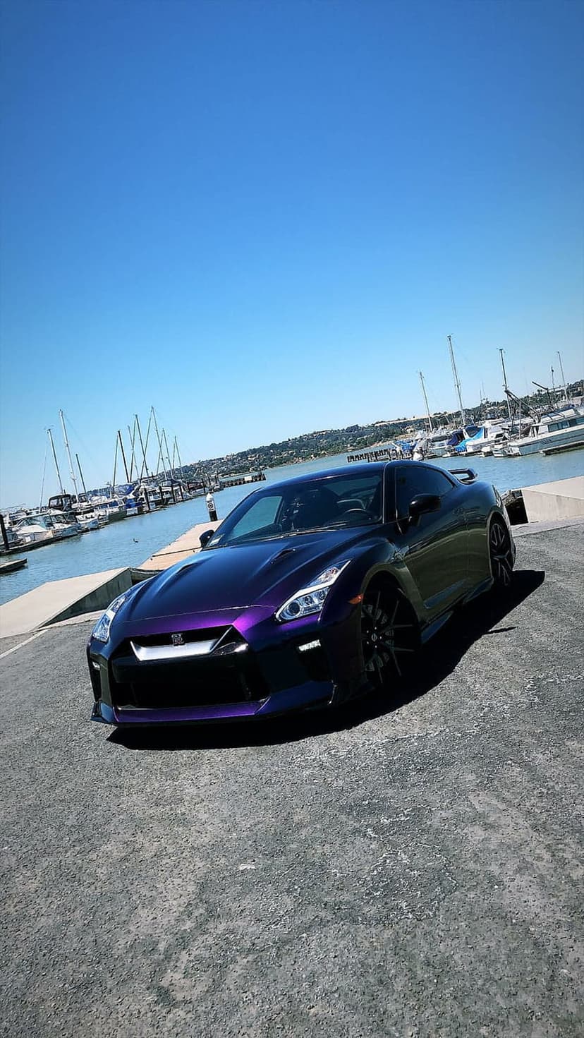 Nissan GT-R in metallic purple parked on a stone pier with sailboats and a clear blue sky in the background.