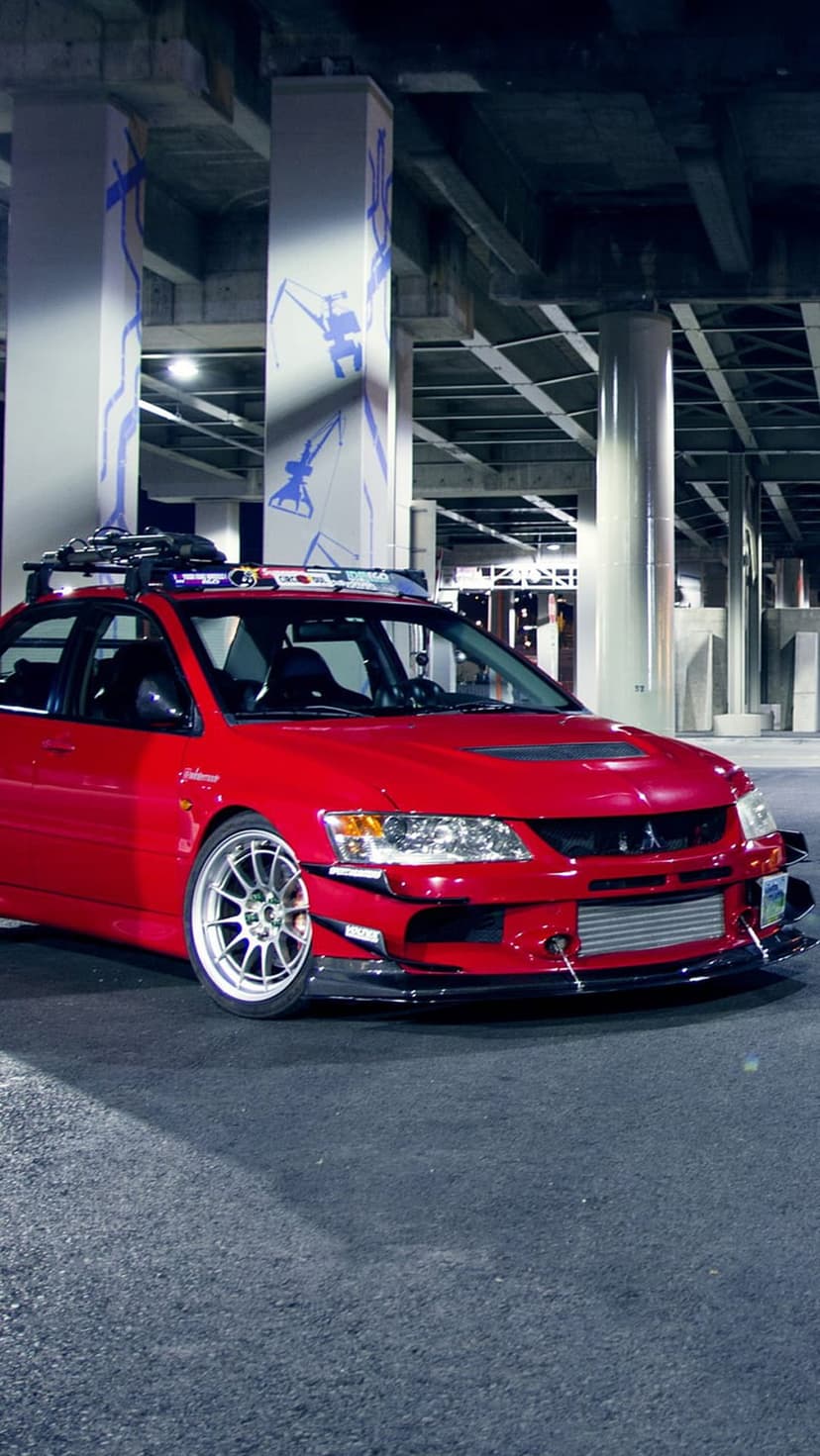 Red Mitsubishi Lancer Evolution parked under concrete pillars with blue crane graffiti art in an urban setting.