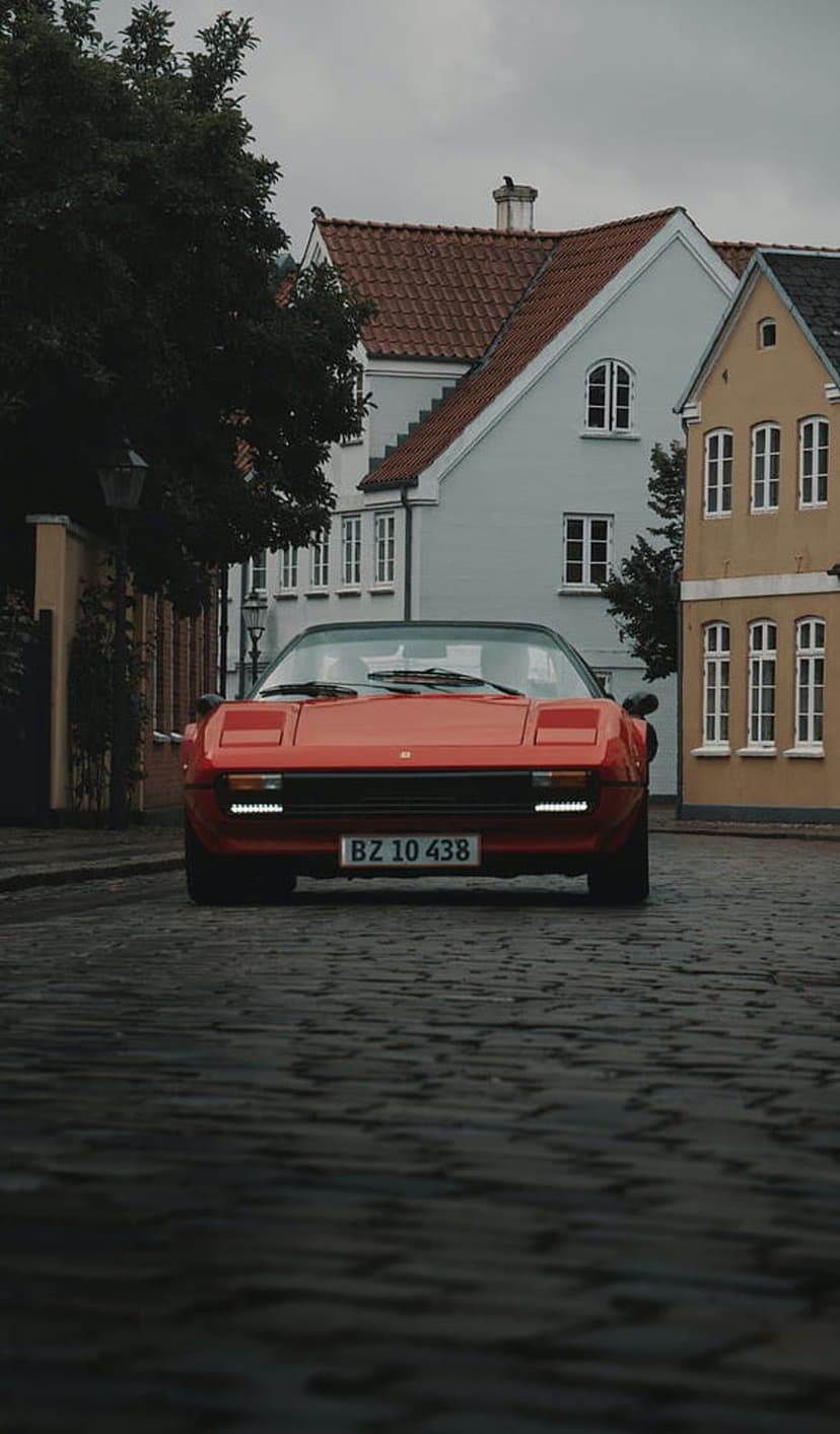 Red vintage Ferrari parked on a cobblestone street between yellow and red European houses under soft light.