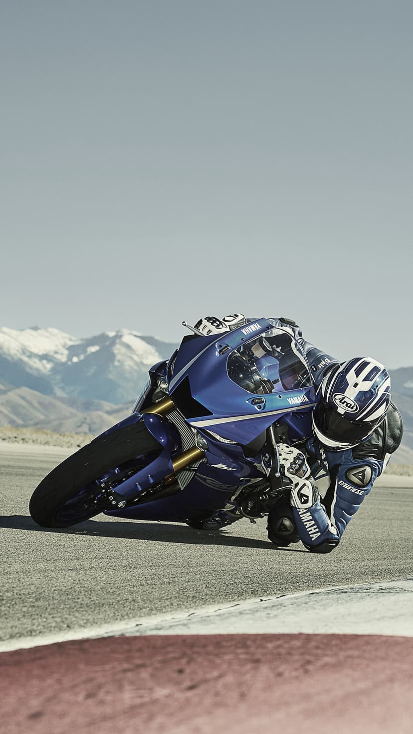 Yamaha motorcycle rider leans a blue sportbike into a track corner with snowy mountain peaks in the distance.