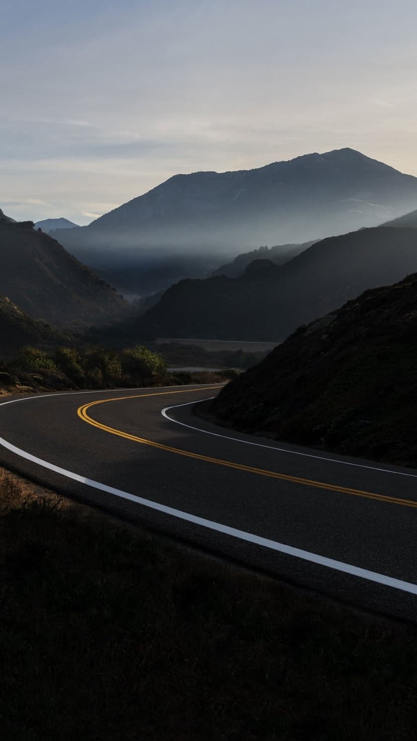 Curving asphalt road with yellow lines winds through hazy blue mountain peaks under soft morning golden sunlight.