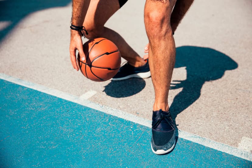 Man's hand and legs dribbling a basketball on a blue and gray court with long shadows under bright sunlight.