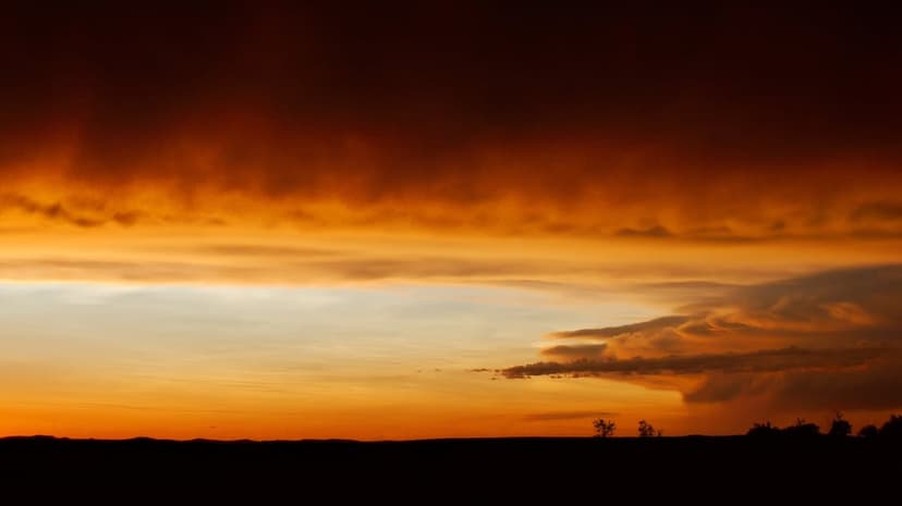 Fiery orange and red clouds swirl above a dark silhouetted horizon line under a pale blue twilight sky.