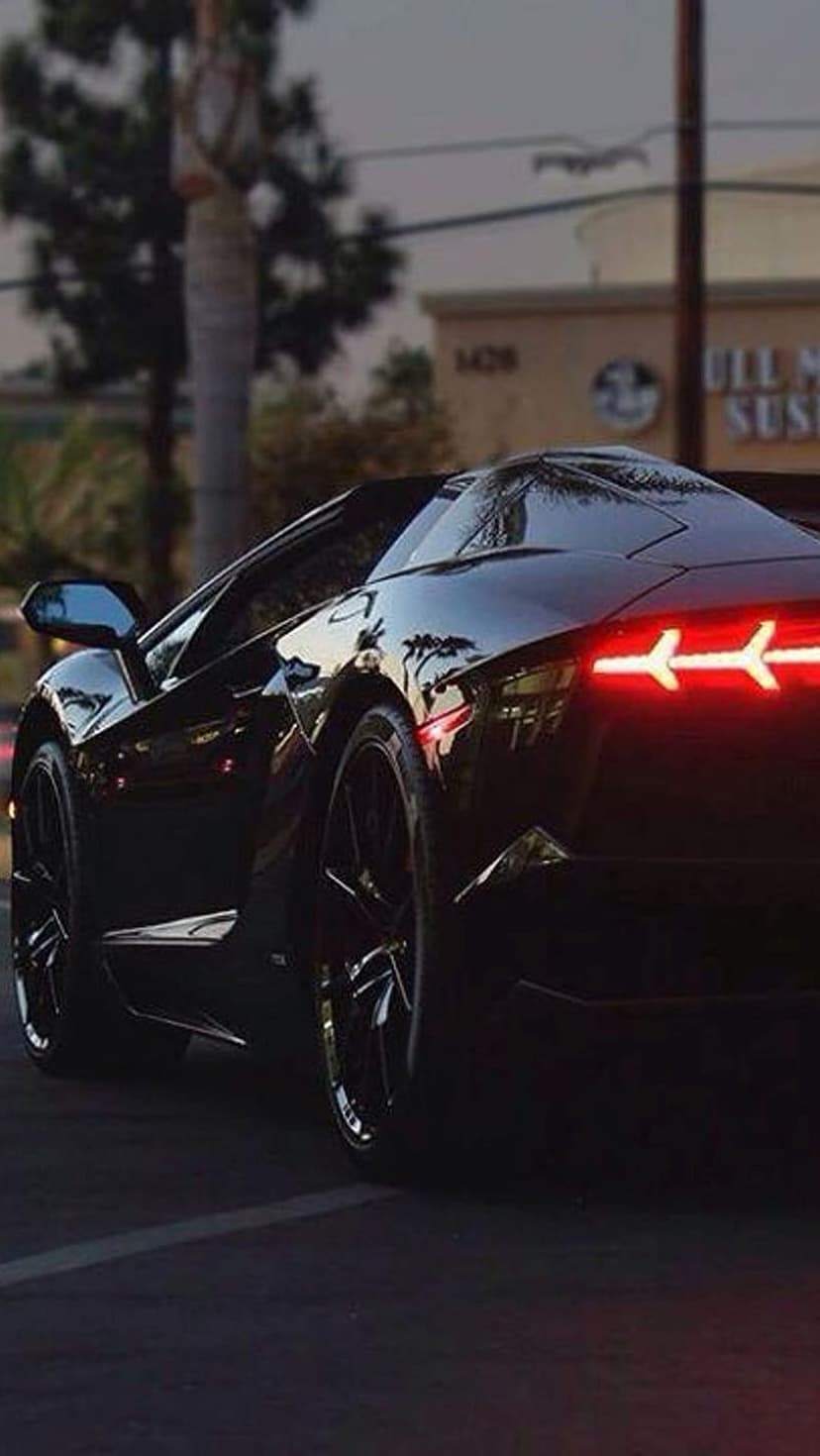 Black Lamborghini Aventador rear view with glowing red Y-shaped LED lights on a city street at twilight.