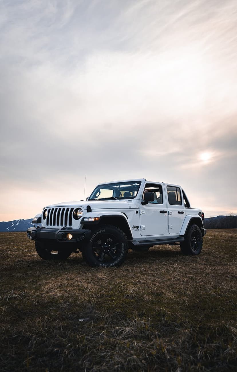 White Jeep Wrangler with black wheels in a dry grassy field before mountains during a vibrant sunset at dusk.