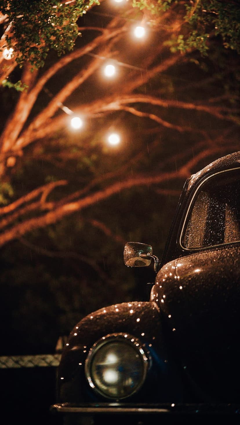 Vintage car headlight and chrome fender bead with rain under warm glowing string lights during a moody rainy night.