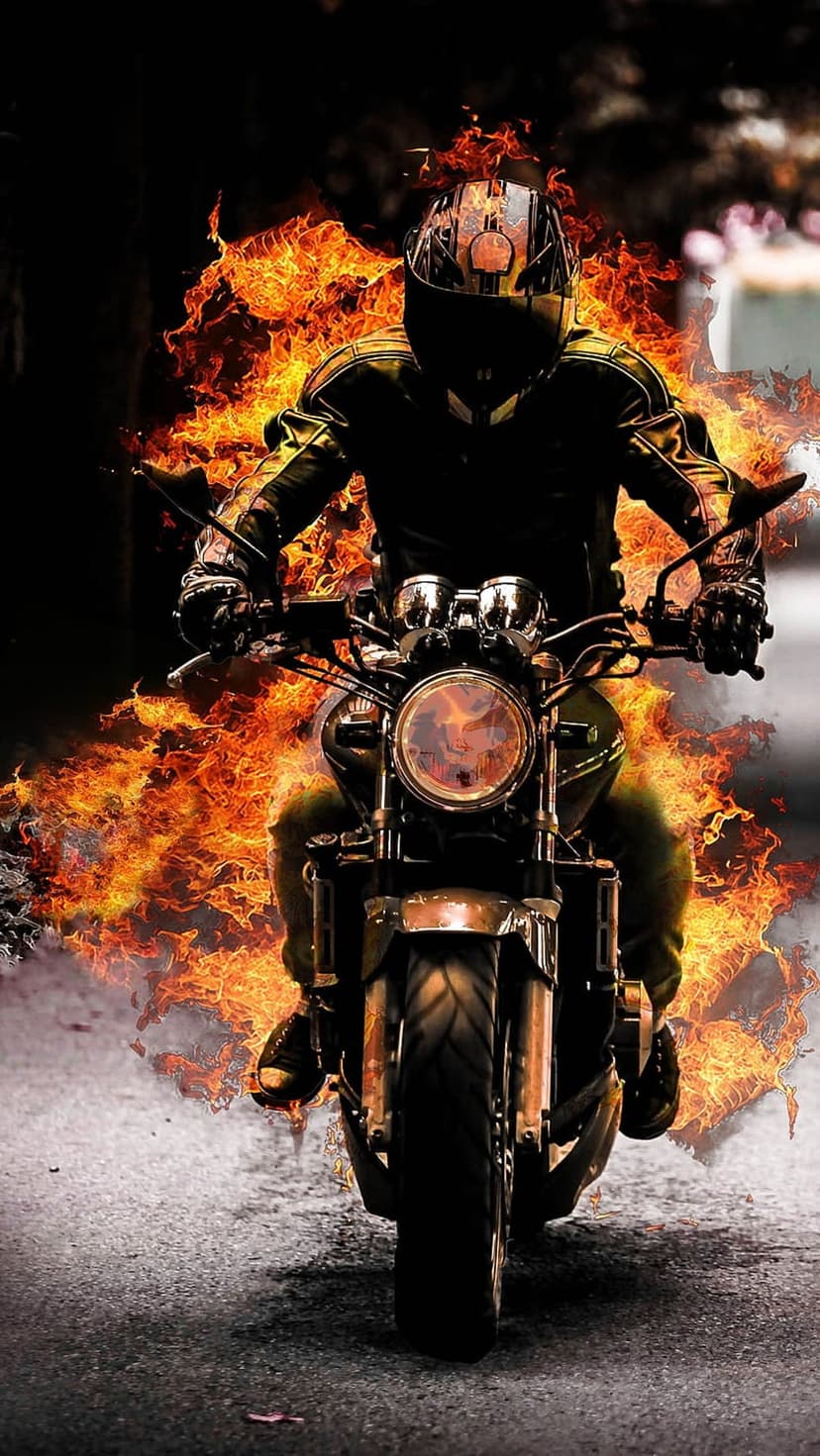 Motorcyclist wearing black gear rides a bike engulfed in orange flames on a dark asphalt road at night.