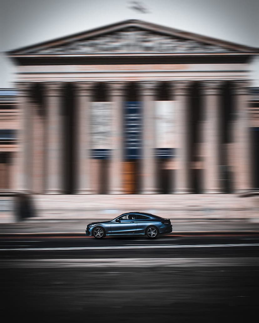Blue coupe car speeds past a classical building with tall columns using a heavy motion blur effect on the road.