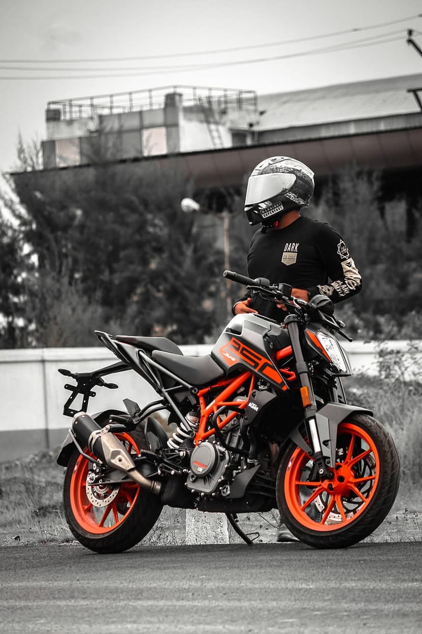 Man in a patterned helmet stands by an orange KTM motorcycle with a desaturated urban building background.