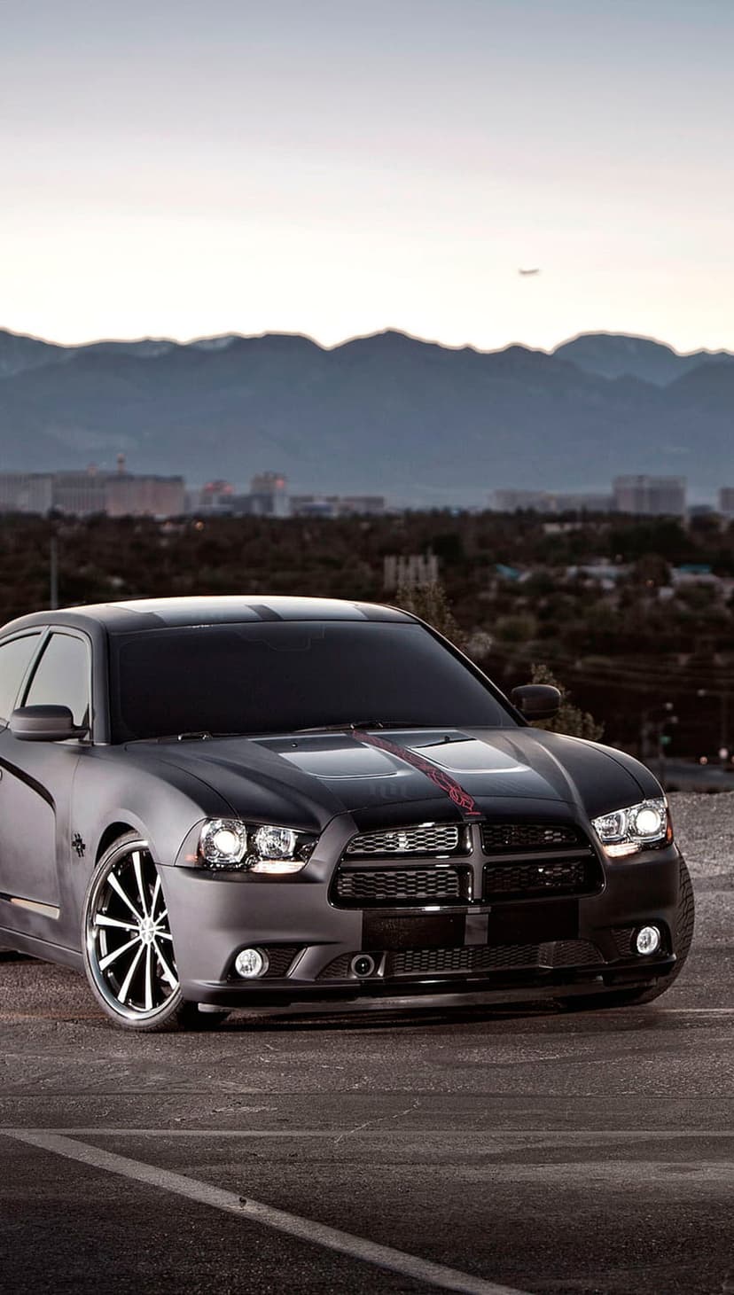 Matte black Dodge Charger with racing stripes on an overlook with a twilight city and distant airplane visible.