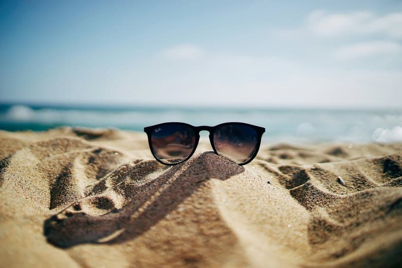 Dark sunglasses resting on rippled beach sand with soft waves and a blue sky blurred in the background.
