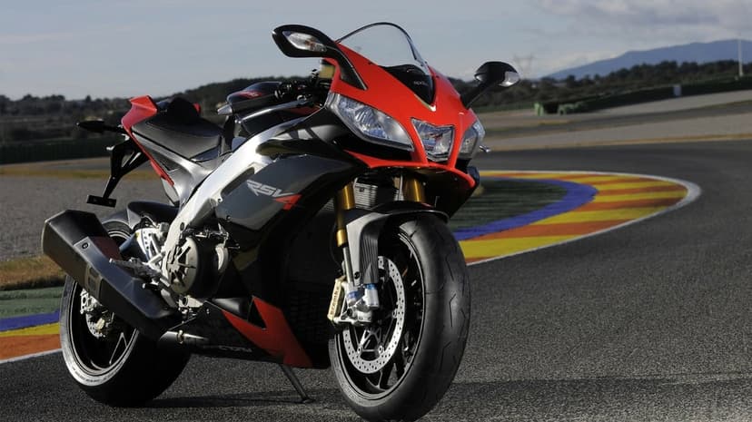 Red and black Aprilia RSV4 superbike parked on a racetrack corner with colorful curbing and distant hills.