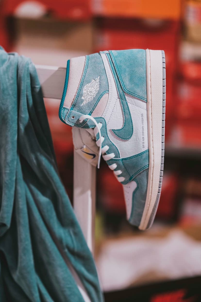 Air Jordan 1 High OG sneaker featuring aqua teal suede and white leather in front of blurred shoeboxes.