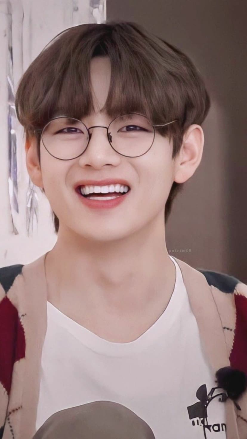 Kim Taehyung of BTS smiles broadly while wearing round glasses and a patterned cardigan in a close-up portrait.