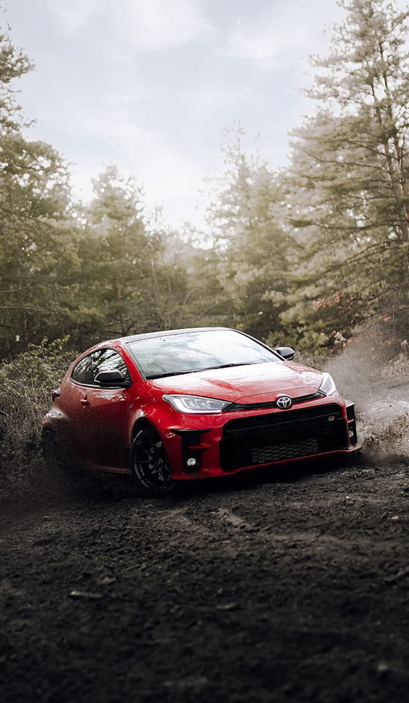 Red Toyota GR Yaris racing through a muddy forest track with dirt kicking up from spinning wheels among tall trees.