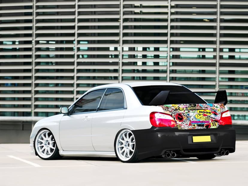 Modified white Subaru WRX STI with a sticker-bomb trunk, carbon fiber bumper, and large rear wing on a plaza.