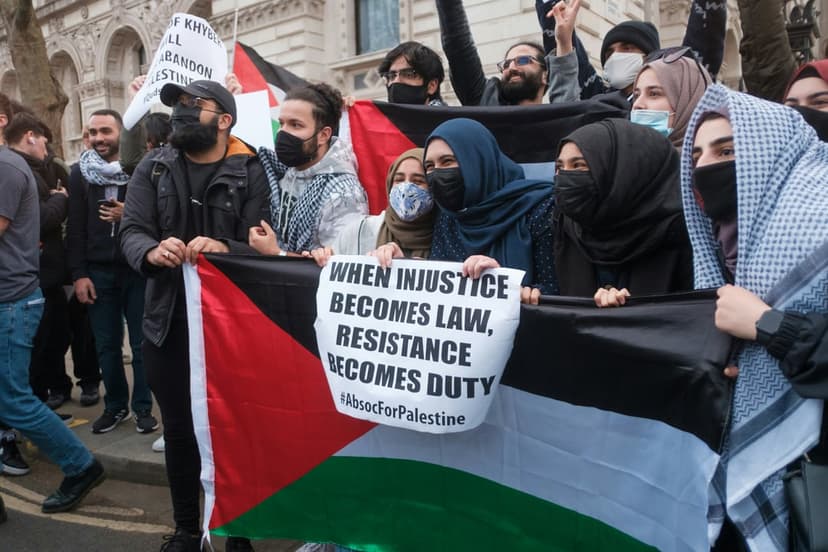 Palestinian flag and resistance banner held by protesters wearing keffiyeh scarves during an outdoor rally.