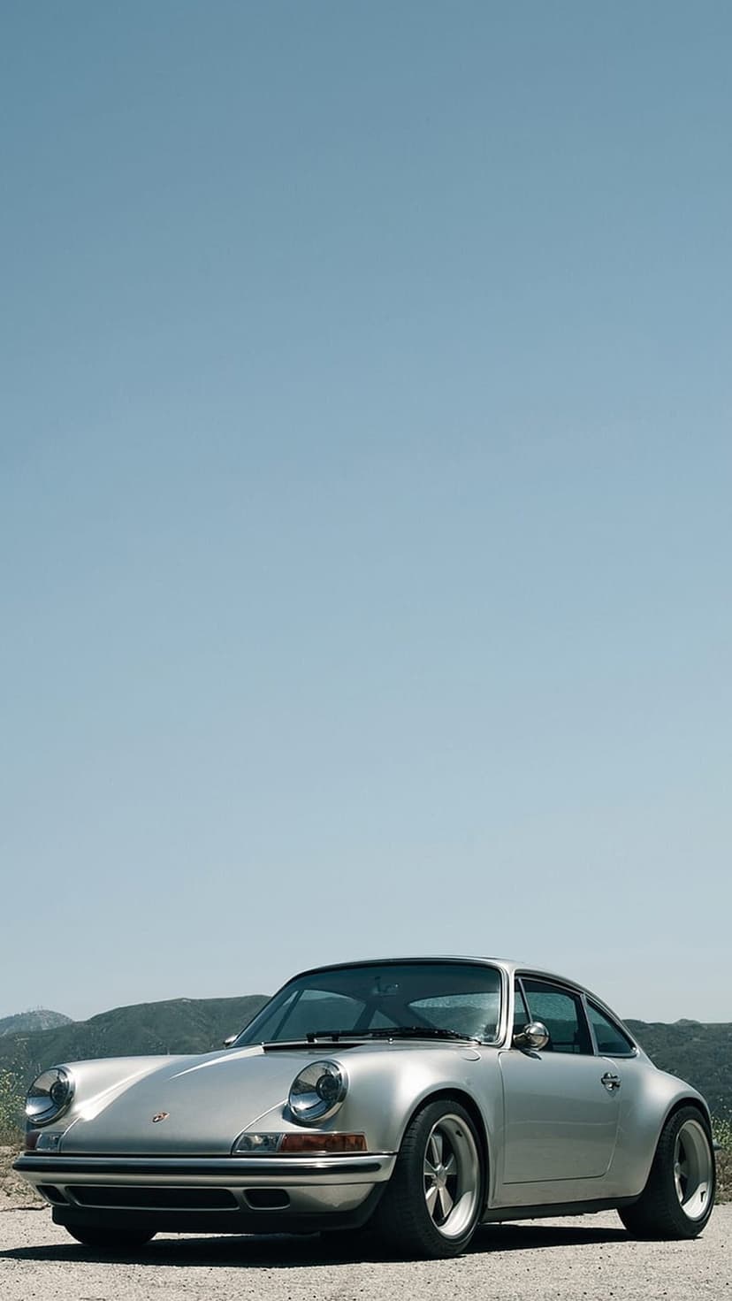 Silver Porsche 911 coupe parked on a gravel trail with jagged mountain peaks under a bright blue sky.