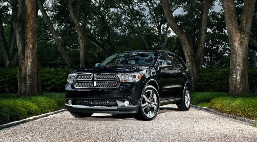 Black Dodge Durango SUV parked on a gravel path under tall green trees with bright daylight reflections.