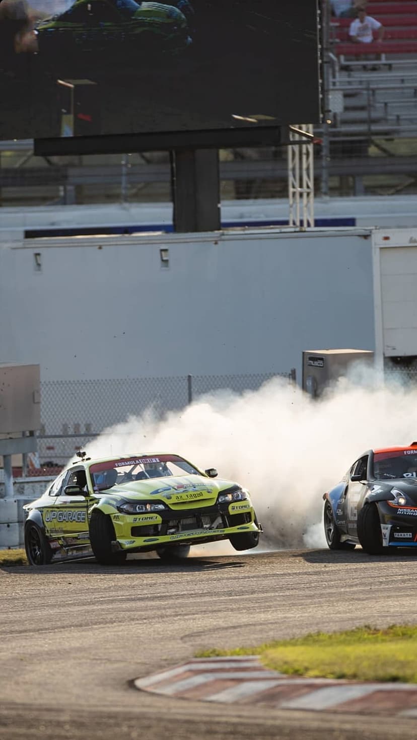 Race cars drift along a track with red and white curbs, producing a large white cloud of smoke in the sunlight.