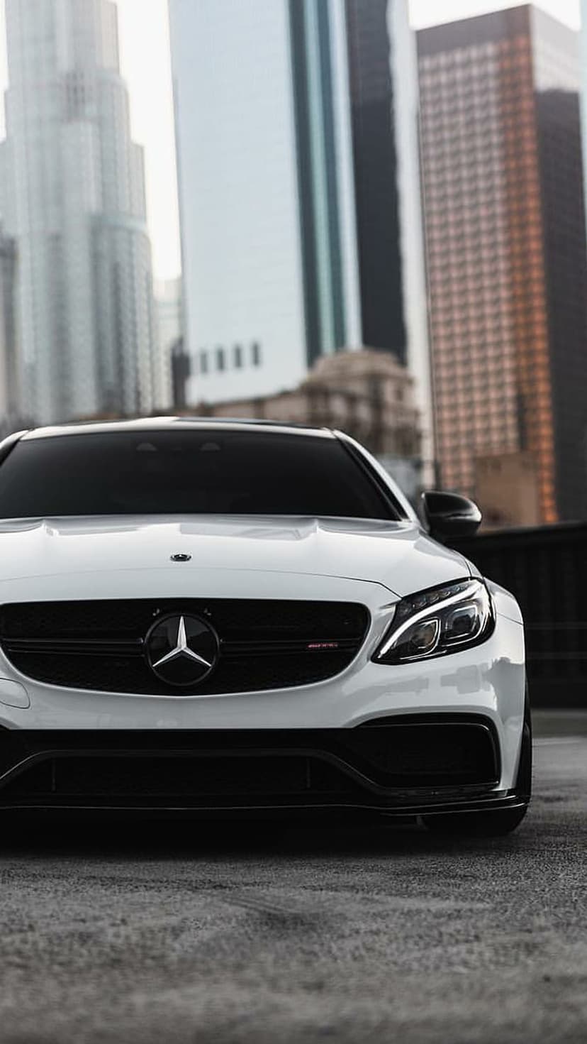 White Mercedes-AMG C63 S Coupe with chrome grille parked on asphalt against a blurred city skyscraper backdrop.