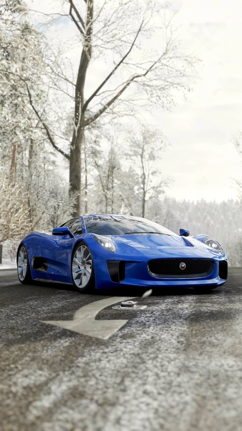 Blue Jaguar sports car driving on a snowy road through a dense forest with frost-covered trees and a gray sky.